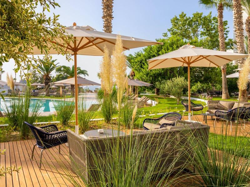 Eine Terrasse mit Holzdeck, Sonnenschirmen, schwarzen Stühlen, Pflanzen und Palmen im Hintergrund.