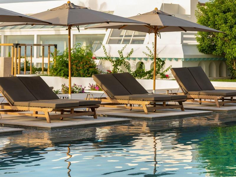 Poolbereich mit braunen Liegestühlen und großen weißen Sonnenschirmen neben modernem Gebäude.