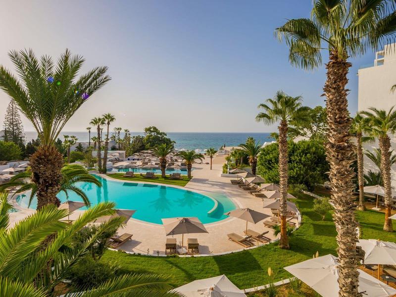 Schwimmbad mit Palmen und Sonnenliegen, Blick aufs Meer und blauer Himmel im Hintergrund.