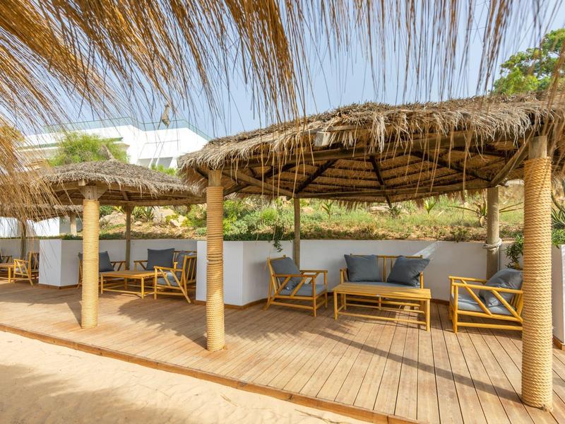 Offener Strandpavillon mit Bambusmöbeln, Strohdach und Holzterrasse an sonnigem Tag.