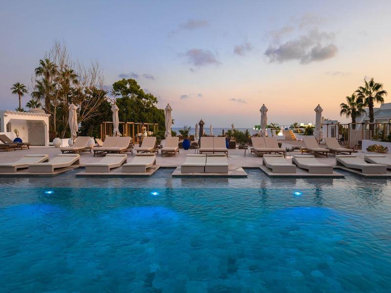 Großer Pool mit blauer Beleuchtung, Liegen und Palmen bei Sonnenuntergang am Meer.
