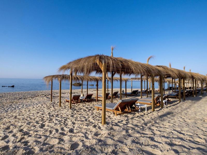Strand mit Liegen und Schilfdächern unter klarem blauem Himmel am ruhigen Meer.