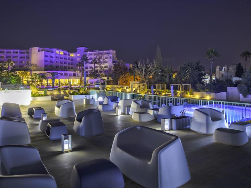 Moderner Außenbereich mit beleuchteten Sitzecken und Pool vor einem mehrstöckigen Hotel bei Nacht.