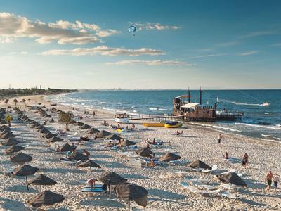 Weißer Sandstrand mit Strohsonnenschirmen und Menschen, daneben ein Gebäude am Meer.