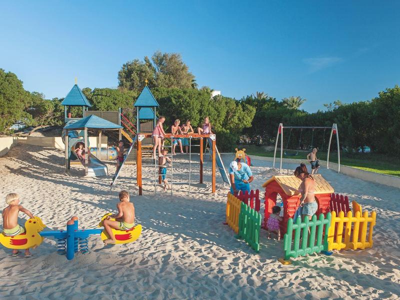 Des enfants jouent dans une aire de jeu de sable avec balançoires et structures d'escalade sous un ciel clair.