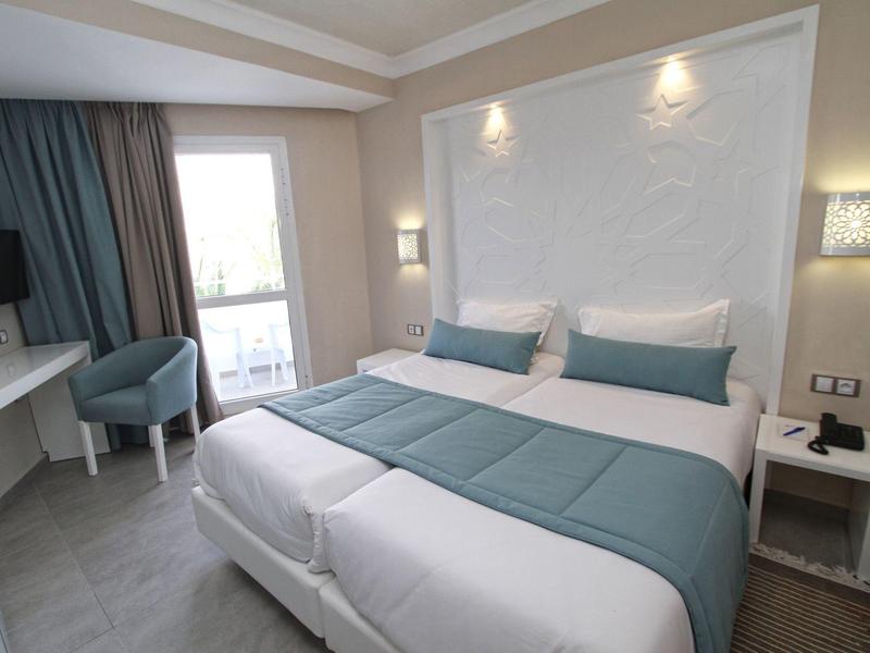 Chambre d'hôtel moderne avec grand lit, bureau, fauteuils et balcon avec rideaux.