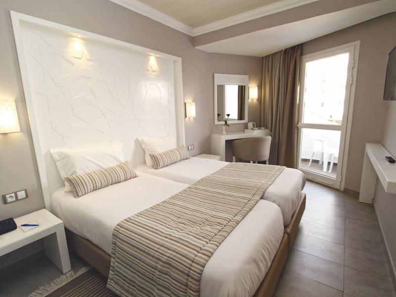 Chambre d'hôtel lumineuse avec deux lits simples, bureau et fenêtre donnant sur balcon.