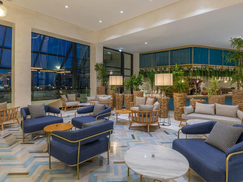 Světlá hotelová hala s moderními modrými křesly a velkými okny s nočním výhledem na město.