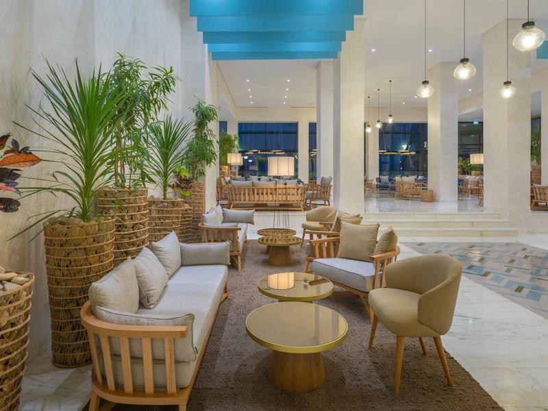 Moderní hotelová lounge oblast s rostlinami, pohovkami a židlemi v světlém prostředí.