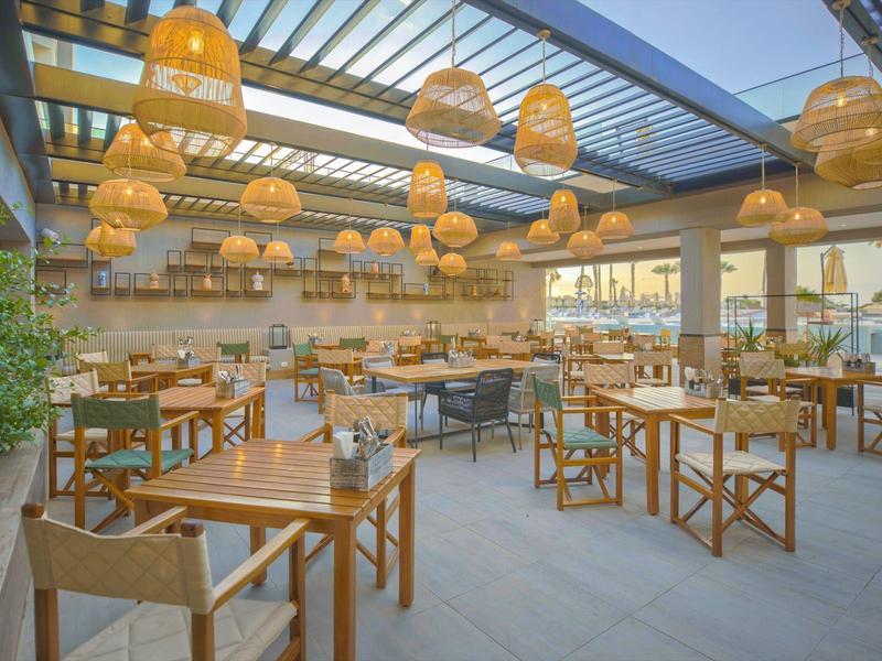 Venkovní restaurace s dřevěným nábytkem a dekorativními lampami pod skleněnou střechou.