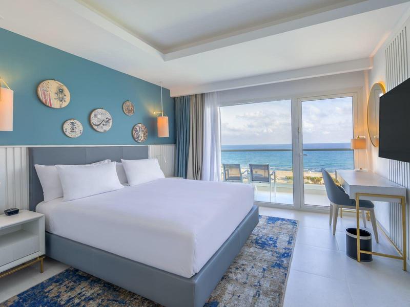 Moderní hotelový pokoj s velkou postelí, výhledem na moře a balkonem za denního světla.