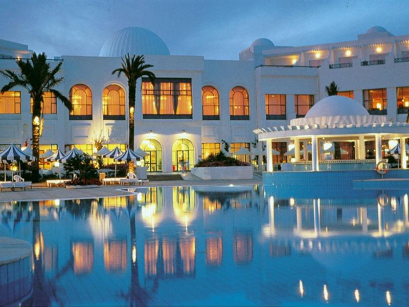 Elegante hotel met zwembad, palmbomen en verlichte ramen bij schemering.