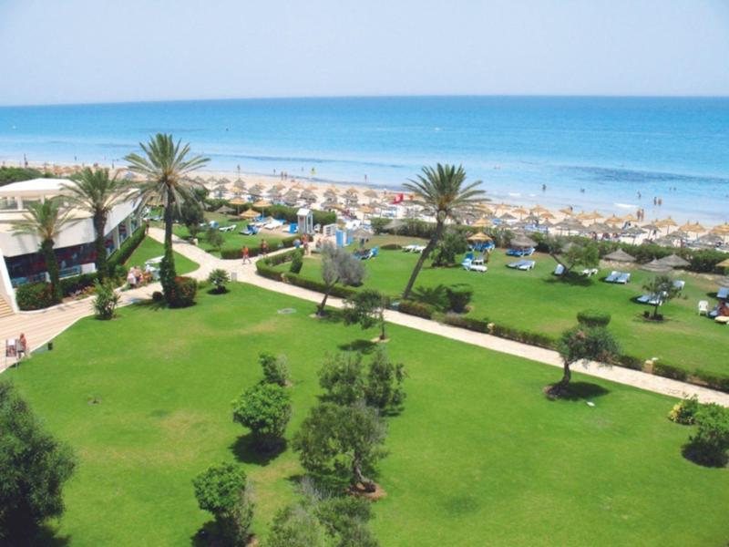 Groene tuin met palmbomen, ligstoelen en uitzicht op het zandstrand en de blauwe zee.