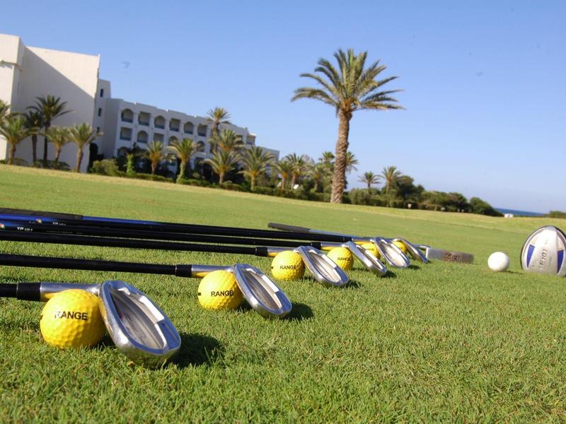 Golfballen en clubs liggen op gras voor een hotelgebouw en palmbomen onder een blauwe lucht.
