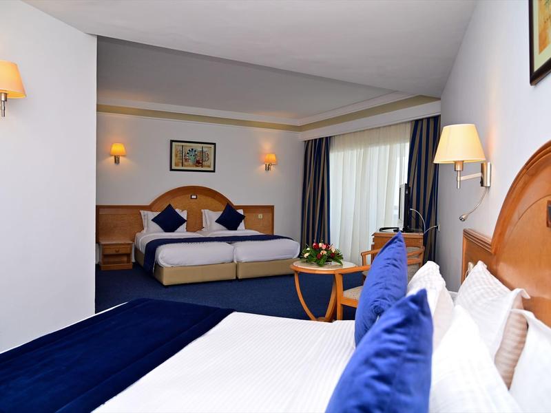 Lichte hotelkamer met twee bedden, blauwe kussens en een kleine tafel.