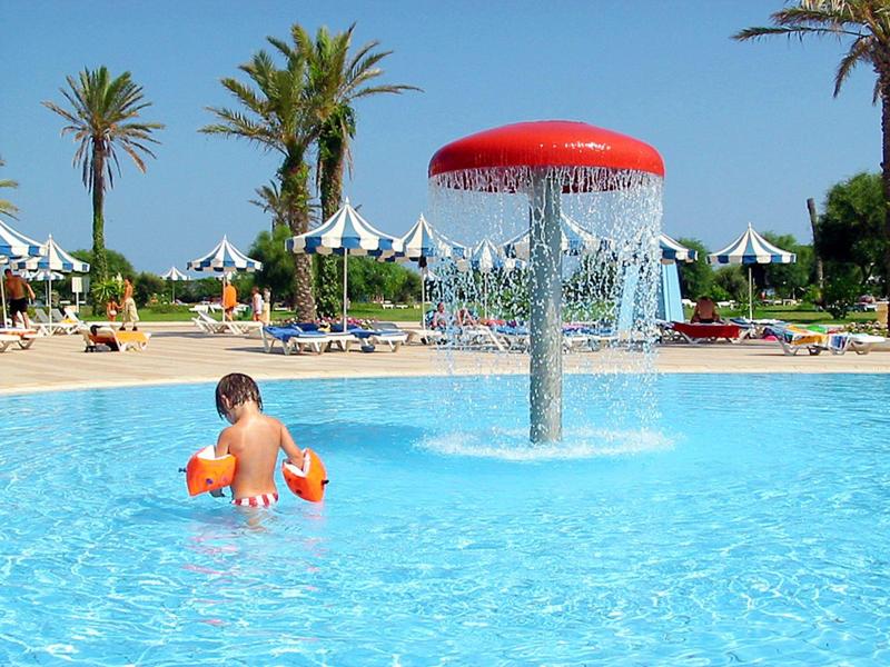 Kind speelt in het zwembad met waterpaddenstoel en palmen op de achtergrond bij een hotelresort.