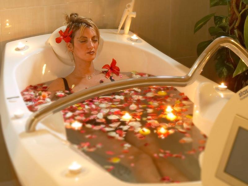 Vrouw ontspant in bad gevuld met rozenblaadjes en kaarslicht in een rustgevende sfeer.