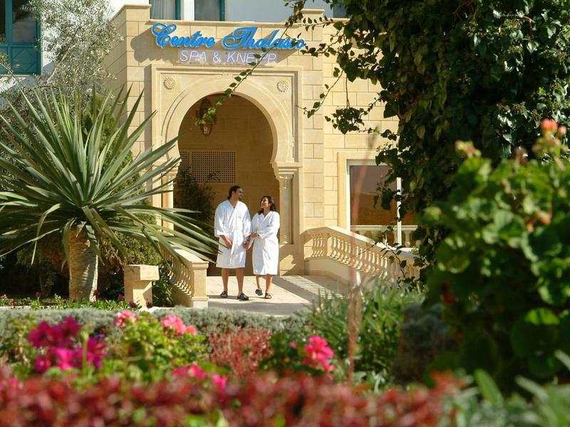 Twee mensen in witte badjassen bij een hotel ingang met planten en bloemen op de voorgrond.