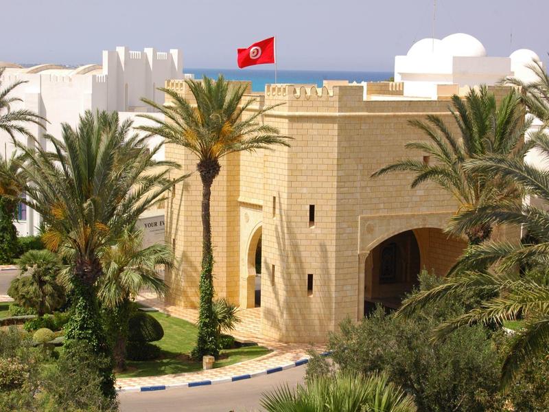 Hotelgebouw met palmentuinen en een rode vlag in een warm zonnig klimaat.