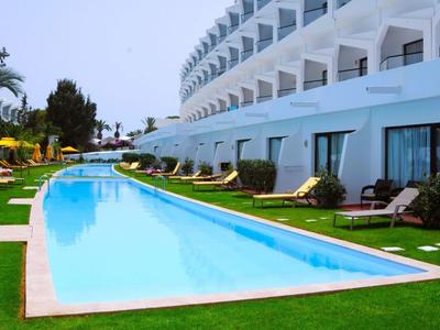 Lang buitenzwembad met ligstoelen voor een meer verdieping tellend hotelgebouw bij heldere lucht.