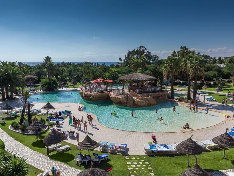 Resort zwembad met ligstoelen, palmbomen en een centrale houten gazebo onder een heldere lucht.