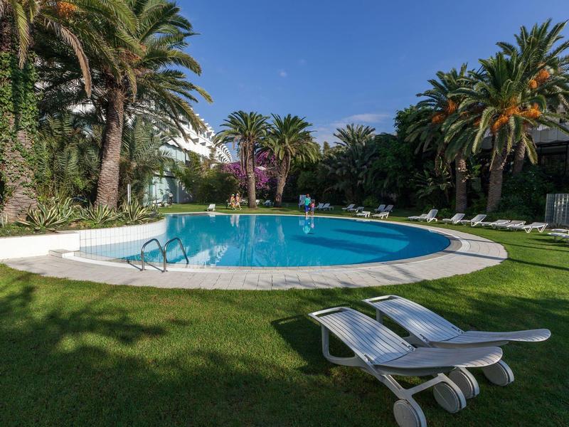 Buitenzwembad omgeven door palmbomen en twee witte ligstoelen op groen gras.