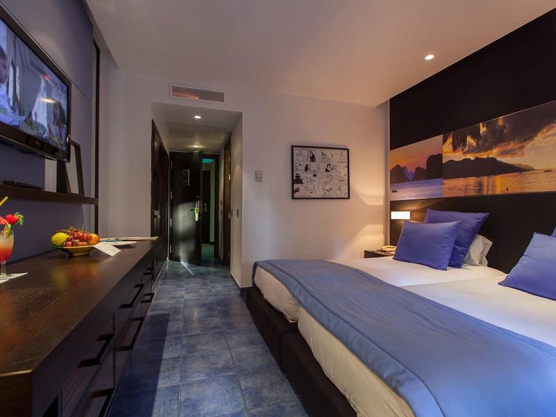 Moderne hotelkamer met tweepersoonsbed, blauwe kussens, tv en zonsondergang muurkunst.