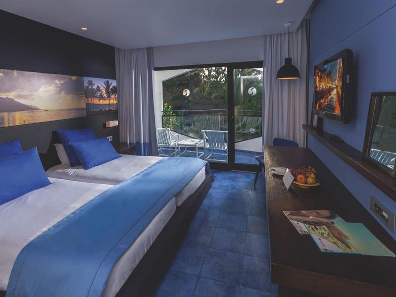 Modern hotelkamer met twee bedden, blauwe muren en uitzicht op een terras met zwembad.