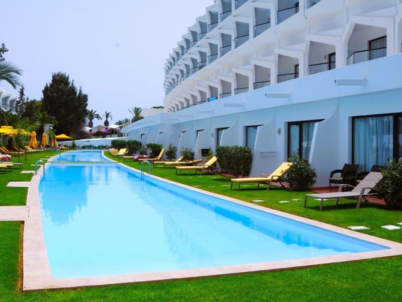 Lang buitenzwembad met ligstoelen voor een meer verdieping tellend hotelgebouw bij heldere lucht.