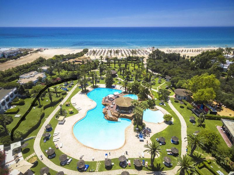 Groot zwembad met palmbomen en ligstoelen bij een zandstrand en blauwe zee.