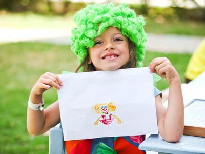 Kind sitzt draußen mit grünem lockigem Hut und hält ein Papier mit einer bunten Zeichnung.