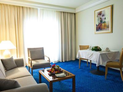 Helles Hotelzimmer mit blauem Teppich, Sofa, Stühlen, Tisch und großem Fenster mit Vorhängen.