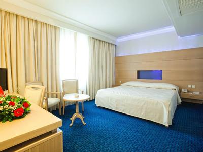 Helles Hotelzimmer mit großem Bett, blauem Teppich und zwei Sesseln vor Fenster mit Vorhängen.