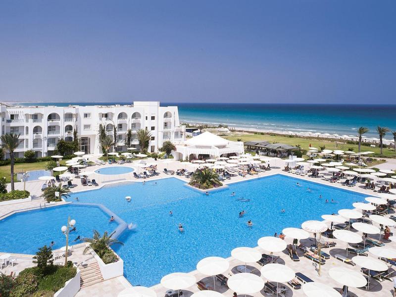 Hotel mit großem Pool, weißen Sonnenschirmen und Blick auf das Meer bei klarem Himmel.