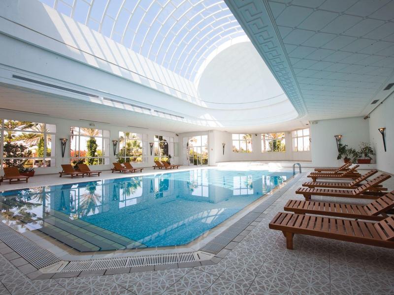Innenpoolbereich mit Sonnenliegen und großer Glasdecke in einem Hotel.