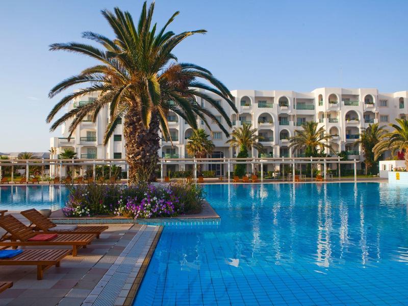 Großer Außenpool mit Liegestühlen und Palmen vor einem mehrstöckigen Hotelgebäude