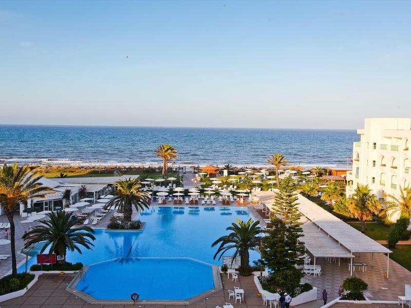 Blick auf einen Hotelpool mit Palmen und Meer im Hintergrund bei klarem Himmel.