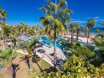 View of a hotel pool with palm trees and sea in the background under a sunny sky.