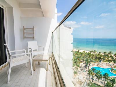Balcon avec chaises blanches et vitrages offrant une vue sur des palmiers et une piscine au bord de la mer.