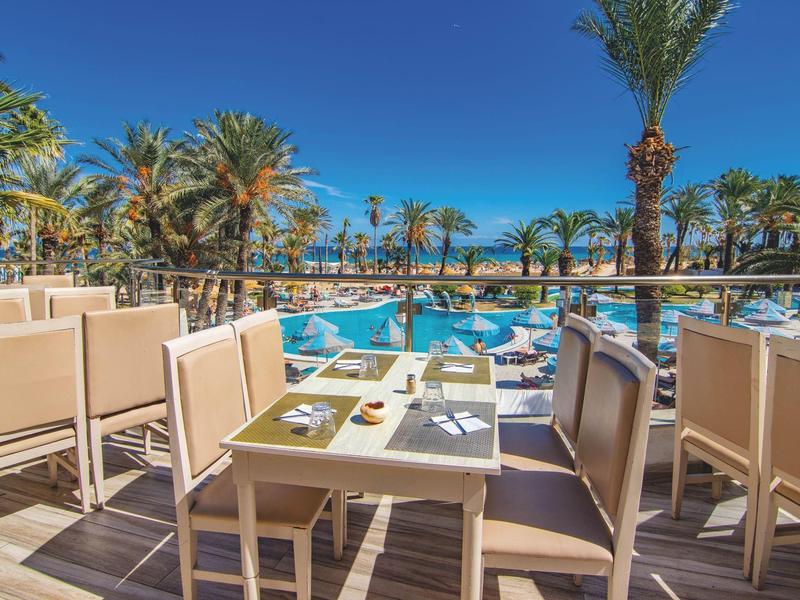 Terrasse avec tables et chaises près d'une piscine et des palmiers sous un ciel clair.