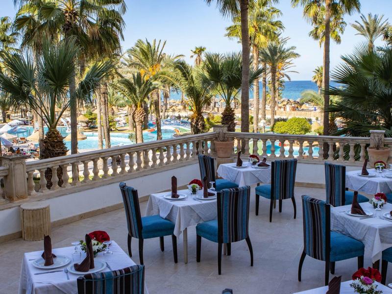 Terrasse avec tables, chaises, palmiers et vue sur la mer dans un hôtel.