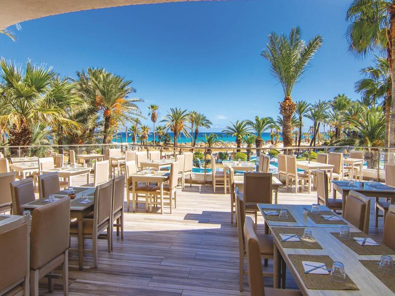 Terrasse de restaurant avec vue sur piscine et palmiers sous un ciel bleu.