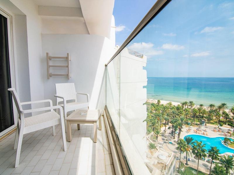 Balcon avec chaises blanches et vitrages offrant une vue sur des palmiers et une piscine au bord de la mer.