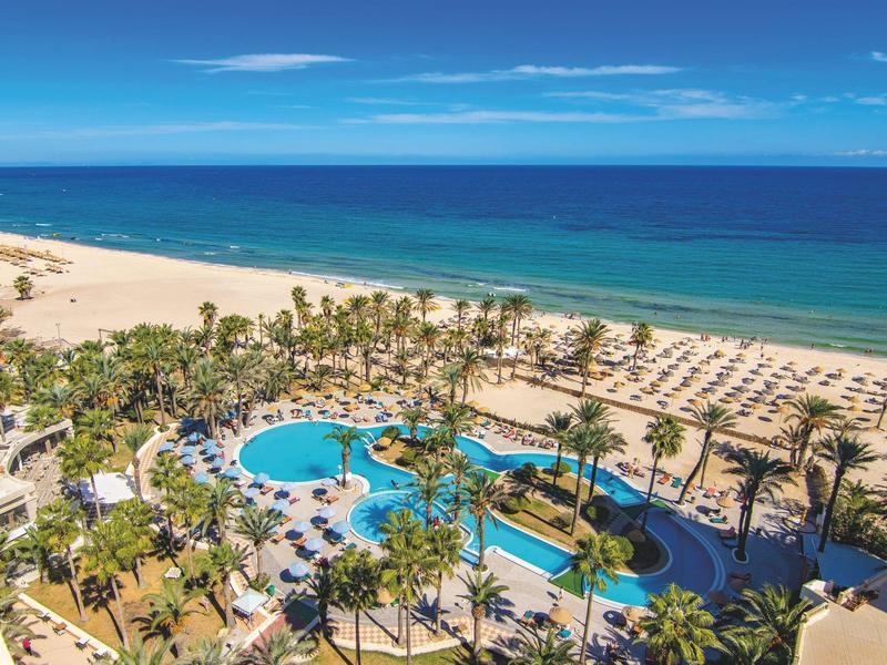 Vue panoramique d'un hôtel avec piscine, palmiers et plage de sable devant une mer bleue.
