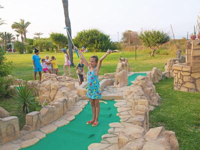 Kinder spielen Minigolf auf einem sonnigen, grünen Platz mit künstlichen Steinen und Palmen.