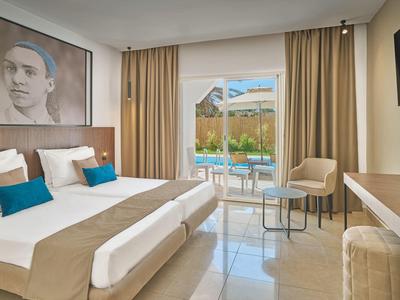 Helles Schlafzimmer mit Doppelbett, cremefarbenen Vorhängen, Stühlen und Blick auf Pool außen.
