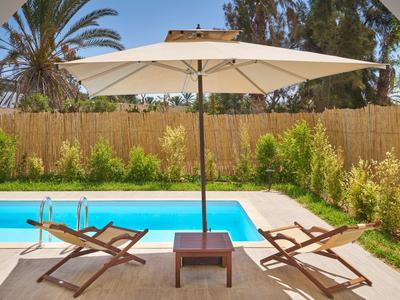 Zwei Holzliegen mit Polstern unter großem weißen Sonnenschirm am blauen Pool im Garten.