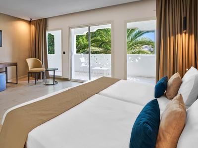 Modernes, helles Hotelzimmer mit großem Bett, Panoramafenstern und Balkon mit grünem Ausblick.