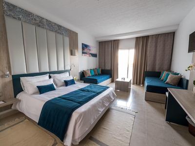 Modern eingerichtetes Hotelzimmer mit großem Bett, Sofas, TV und Balkon mit Vorhängen.