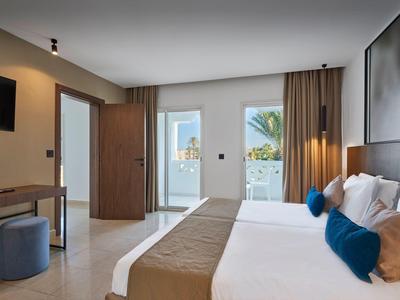Modernes Hotelzimmer mit Doppelbett, weißen Wänden, braunen Vorhängen und Balkon mit Meerblick.